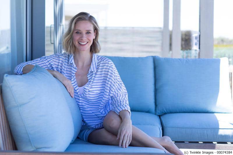 Blonde Frau im gestreiften Hemd und weiflen Shorts sitzt auf einer Couch